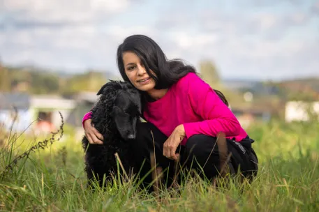 Eine Frau in einem pinken Shirt kniet mit ihrem schwarzen Hund auf dem Gras.