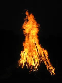 Ein großes Lagerfeuer brennt hell in der dunklen Nacht und wirft ein warmes Licht auf die Umgebung. Die Flammen sind intensiv und reichen hoch in die Luft.