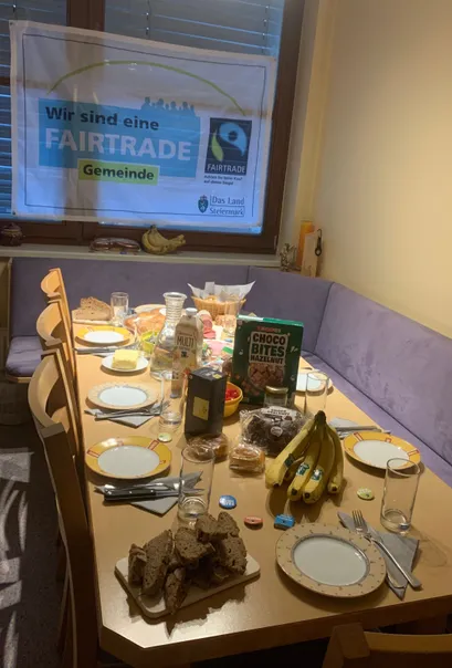 Ein Esstisch ist mit Tellern, Gläsern und Essen gedeckt. Eine Box Choco Bites Haselnüsse und Bananen stehen auf dem Tisch. Ein Schild steht Wir sind eine FAIRTRADE Gemeinde.