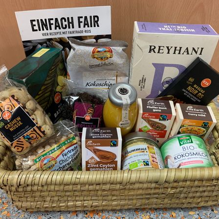 Ein Korb mit verschiedenen Bio- und Fair-Trade-Lebensmitteln auf einer Arbeitsplatte, darunter Hummus, Müsli, Nüsse und ein Schild, das Einfach Fair sagt.