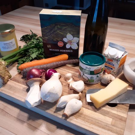 Ein Holztisch mit einem Schneidebrett hat Pilze, Karotten, Rüben, Zwiebeln, Knoblauch, Käse, eine Flasche Wein, eine Schüssel und eine Packung Jasminreis.