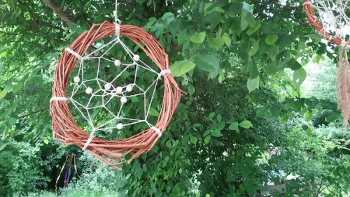 Ein Traumfänger aus braunem Draht hängt von einem Ast, umgeben von grünen Blättern. Er hat eine spinnennetzartige Struktur mit weißen Perlen.
