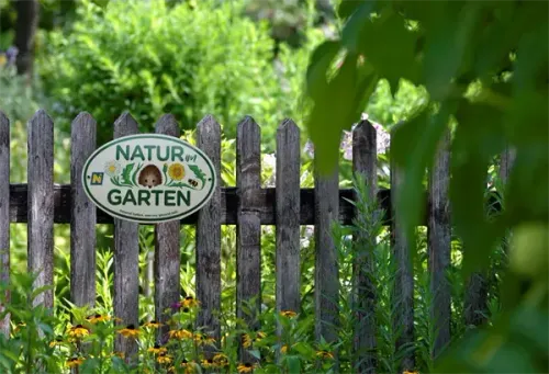 Ein Holzzaun mit einem Schild, das 'Natur am Garten' besagt, ist von verschiedenen Pflanzen und Blumen umgeben.