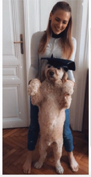 Eine Frau, die einen Hund mit einer Doktormütze hält und lächelt, steht drinnen mit einem Holzboden und einer weißen Tür hinter ihr.