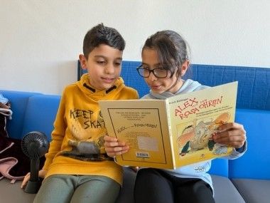 Zwei Kinder sitzen auf einer blauen Couch und lesen ein Buch mit dem Titel Alex Rosa Chicken. Der Junge trägt eine gelbe Hoodiejacke und das Mädchen eine Brille.