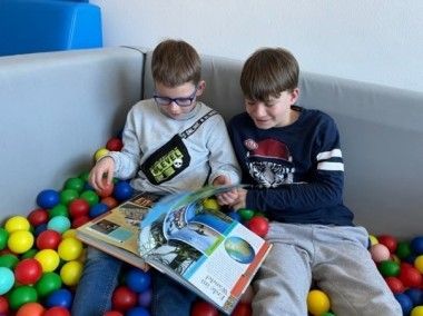 Zwei Jungen mit Brille sitzen auf einer Couch voller bunter Bälle. Einer hält ein offenes Buch, während der andere ihn ansieht. Die Wand hinter ihnen ist weiß.