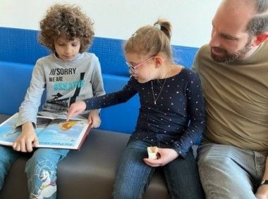 Ein Junge und ein Mädchen sitzen auf einer blauen Couch und schauen in ein Buch. Der Junge zeigt auf eine Seite und das Mädchen trägt eine Brille. Ein erwachsener Mann sitzt neben ihnen.
