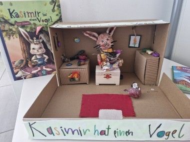 Ein Karton-Diorama zeigt einen kleinen Raum mit einer Hasenfigur, einem kleinen Tisch und einem roten Teppich. Es gibt zwei kleine Schubladen und ein Buch mit dem Titel 'Kasimir hat ein Vogel'.