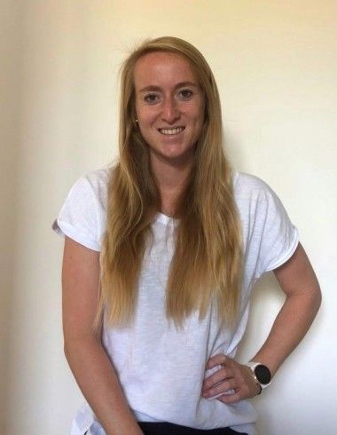 Eine Frau mit langen blonden Haaren steht und lächelt für ein Foto. Sie trägt ein weißes Shirt und schwarze Hose, und ihre Hand liegt auf der Hüfte.