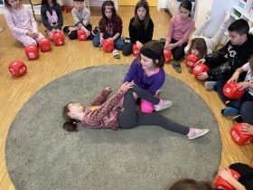 Kinder sitzen im Kreis auf dem Boden mit roten Boxhandschuhen um sie herum, ein Mädchen liegt auf dem Boden und ein anderes hilft ihr beim Aufstehen.