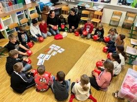 Eine Gruppe von Kindern, die rote Boxhandschuhe tragen, sitzen im Kreis auf einem Holzboden und halten Karten mit Buchstaben. Ein Erwachsener steht hinter ihnen, möglicherweise im Unterricht.