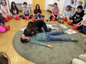 Kinder sitzen im Kreis auf dem Boden, einige tragen rote Boxhandschuhe. Ein Kind liegt auf dem Boden, ein anderes hilft. Eine Lehrerin hilft dem Kind auf dem Boden.