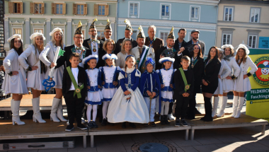 Eine Gruppe von Menschen in passenden weißen und blauen Kostümen steht auf einer Bühne und lächelt für ein Foto. Dahinter sind Gebäude mit Balkonen und Fenstern zu sehen.
