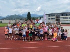 Eine Gruppe von Kindern und Erwachsenen posiert für ein Foto auf einer Laufbahn, mit Bergen und einem Gebäude im Hintergrund.