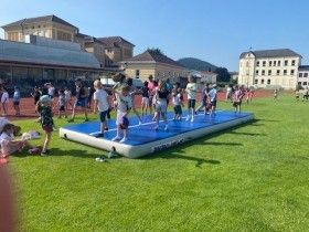 Kinder spielen auf einer blauen aufblasbaren Matte in einem Grasfeld mit Gebäuden im Hintergrund.