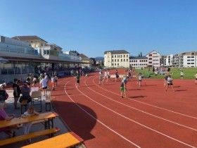 Ein Leichtathletik-Wettkampf an einem sonnigen Tag. Viele Athleten laufen auf der Bahn, Zuschauer sind an der Seite. Im Hintergrund sind Gebäude zu sehen.