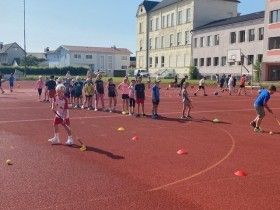 Kinder üben auf einer roten Laufbahn mit Kegeln und Hockeyschlägern. Im Hintergrund befinden sich Gebäude und ein Basketballkorb.