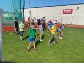 Eine Gruppe von Kindern und Erwachsenen spielt auf einem grünen Feld. Sie tragen Mützen und Turnschuhe. Hinter ihnen steht ein Gebäude mit einem Schild, das 'PAGRO DESIGN' liest.