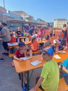 Kinder sitzen an Tischen und essen bei einer Outdoor-Veranstaltung. Einige tragen Hüte und Turnschuhe. Dahinter gehen und sitzen Leute auf Bänken, mit Gebäuden und einer Laufbahn im Hintergrund.