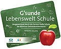 Eine grüne Karte mit dem Text 'G'sunde Lebenswelt Schule' und einem roten Apfel rechts. Die Karte zeigt grüne und blaue Symbole sowie ein weißes Apfelsymbol links.