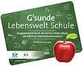 Eine grüne Karte mit dem Text 'G'sunde Lebenswelt Schule' und einem roten Apfel rechts. Die Karte zeigt grüne und blaue Symbole sowie ein weißes Apfelsymbol links.