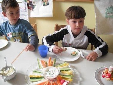 Zwei Jungen sitzen an einem Tisch, einer isst ein Sandwich und der andere eine Karottenstange. Sie haben Teller, Becher und Essen vor sich.