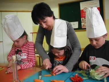 Eine Frau bringt drei Kindern das Schneiden von Gemüse bei. Ein Kind benutzt ein Messer, und die anderen beobachten. Alle tragen Papierkochmützen.