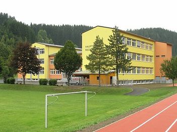 Ein gelbes und oranges Schulgebäude mit vielen Fenstern im Hintergrund, einem Rasenfeld und einer roten Laufbahn im Vordergrund. Bäume und Büsche sind ebenfalls vorhanden.