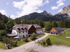 Luftaufnahme eines Berghotels mit einem großen Gebäude und einem kleineren, umgeben von Bäumen und einem Rasenfeld unter einem blauen Himmel mit Wolken.