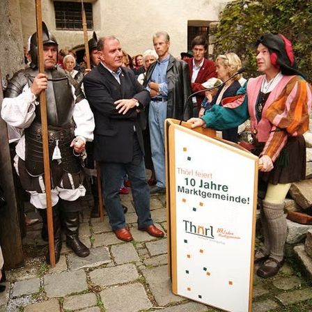 Mehrere Menschen in mittelalterlichen Kostümen stehen auf einem Kopfsteinpflasterweg. Ein Schild zeigt Thorl feiert 10 Jahre Marktgemeinde.
