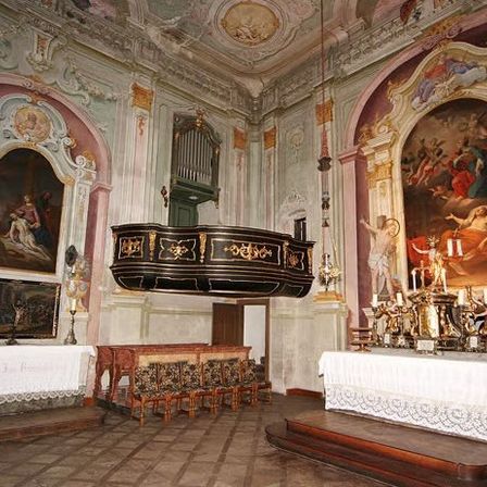 Ein kunstvoll verzierter Kircheninnenraum mit Kanzel, Altar und Gemälden an den Wänden. Kerzen und Skulpturen schmücken den Altar.