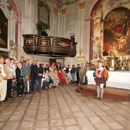 Menschen stehen in einer Reihe vor einem Altar, an dem eine Frau in traditioneller Kleidung steht. Kunstwerke schmücken die Wände, und einige Menschen sitzen auf Bänken.