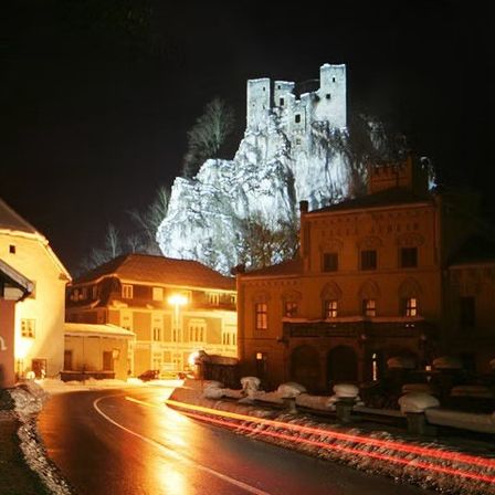 Eine Burg steht nachts auf einem verschneiten Felsen. Die Straße ist leer, und die Straßenlaternen sind eingeschaltet. Schnee liegt auf dem Boden und auf den Bäumen.
