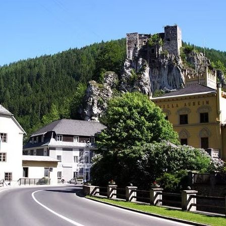 Eine Straße mit einem Weg und mehreren Gebäuden. Die Straße ist leer, mit einem gebogenen Pfad und einer weißen Linie. Bäume und Pflanzen umgeben das Gebiet. Ein Schloss steht auf dem Berggipfel.