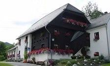Ein weißes und schwarzes Haus mit Blumen in den Fenstern und auf dem Balkon, umgeben von Pflanzen und Blumen, mit Bergen im Hintergrund.