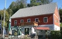 Ein rot-weißes Gebäude mit grauem Dach hat einen Balkon und eine rote Überdachung, umgeben von Bäumen und Pflanzen. Ein Fahnenmast steht links.