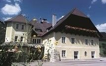Ein großes, cremefarbenes Haus mit einem braunen Dach und mehreren Fenstern steht inmitten von Grün und Bergen im Hintergrund.
