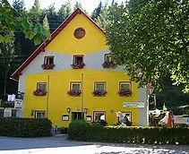 Ein hellgelbes Gebäude mit rotem Rand steht in einem ländlichen Gebiet, umgeben von Bäumen und Sträuchern. Das Haus hat mehrere Fenster mit Blumenkästen und ein rundes Fenster auf dem Giebel.