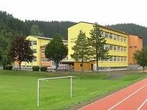 Ein gelbes und orangefarbenes Gebäude mit mehreren Fenstern steht neben einem Grasfeld. Bäume und eine Rennbahn befinden sich vor dem Gebäude.