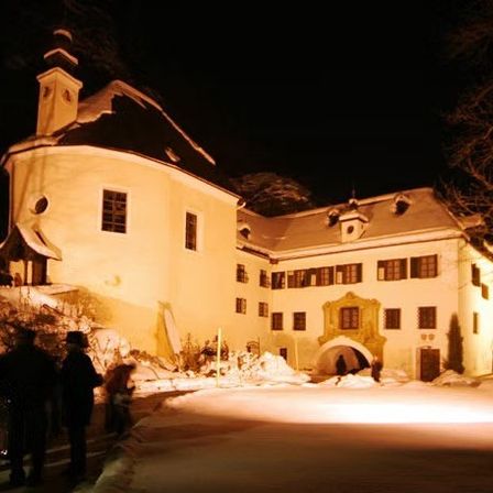 Ein großes beleuchtetes Gebäude bei Nacht mit Menschen davor. Das Gebäude hat viele Fenster und einen Turm.