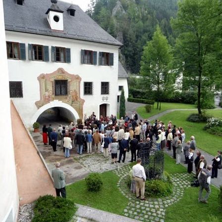 Eine Gruppe von Menschen steht vor einem alten, weißen Gebäude mit einem grauen Dach. Das Gebäude hat einen großen gewölbten Eingang. Die Menschen befinden sich in verschiedenen Positionen und scheinen in ein Gespräch vertieft zu sein. Es gibt Bäume und Büsche in der Umgebung.