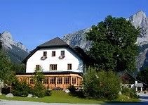 Ein großes weißes Haus mit brauner Einfassung und schwarzem Dach, vor einer Kulisse majestätischer Berge, verfügt über eine große Veranda und mehrere Fenster.