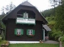 Ein rustikales, schwarzes Chalet mit grünen Fensterläden und einem Balkon, umgeben von üppigem Grün und Bäumen. Das Haus befindet sich in einem bewaldeten Gebiet.