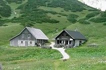 Zwei rustikale Hütten liegen in einem üppigen grünen Tal, verbunden durch einen kleinen Kiesweg. Der Himmel ist klar, und die Landschaft ist mit Wildblumen gespickt.