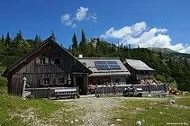 Ein Holzhaus mit Solarpanelen auf dem Dach, umgeben von üppigem Grün und Bergen. Vorne steht eine Bank und ein Zaun.