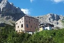 Ein modernes Gebäude mit Glasfenstern steht auf einem Hügel, umgeben von üppigem Grün, mit majestätischen Bergen im Hintergrund unter einem klaren blauen Himmel.