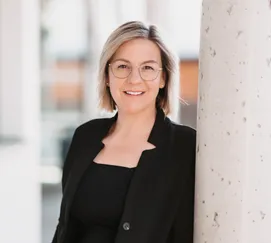 Eine Frau mit kurzen blonden Haaren, die eine Brille und einen schwarzen Blazer trägt, lächelt für ein Foto in einer modernen Büroumgebung.