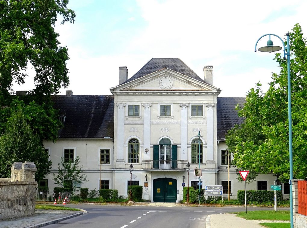  Abschnitt Bild: Ein großes weißes Gebäude mit einer Uhr über dem Eingang hat grüne Fensterläden und eine grüne Tür. Es ist von Bäumen und Büschen umgeben.