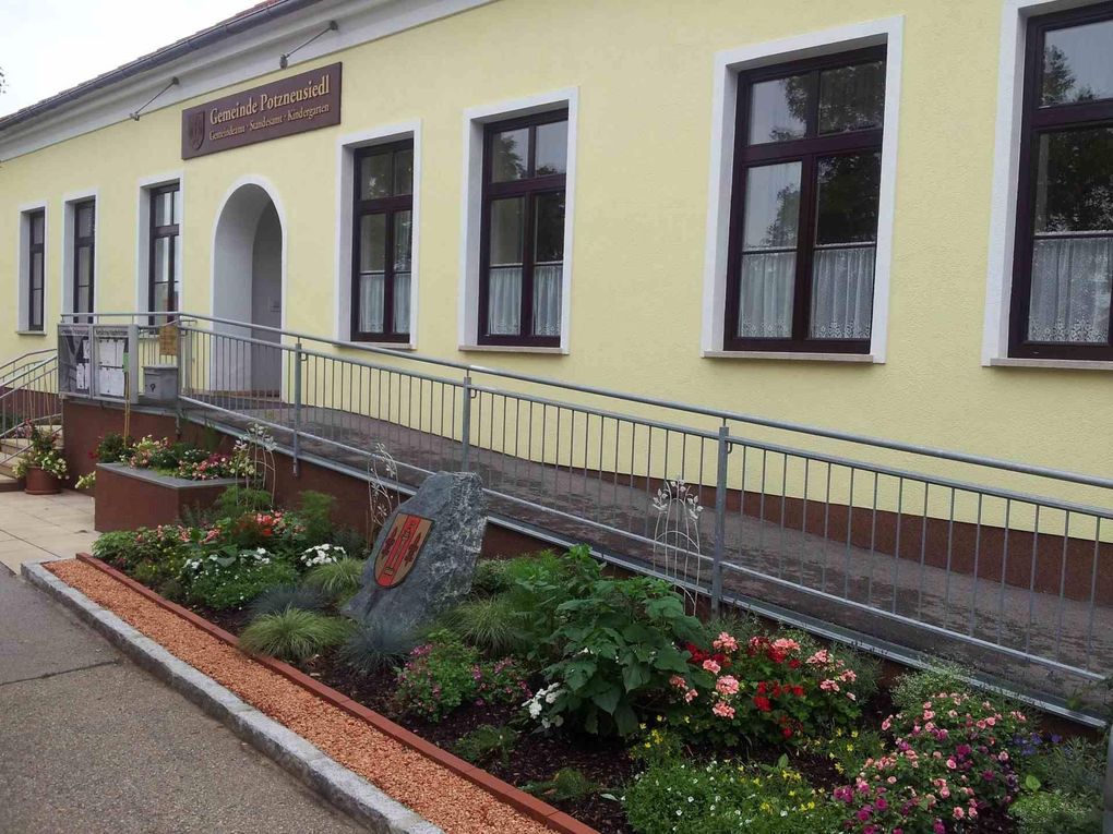 Abschnitt Bild: Ein gelbes Gebäude mit weißem Rand hat drei Fenster und ein Schild mit der Aufschrift 'Gemeinde Porzneusiedl'. Vor dem Gebäude befindet sich ein Metallgeländer, und neben dem Gebäude steht ein Garten mit Blumen und einem Stein mit einem Wappenschild.