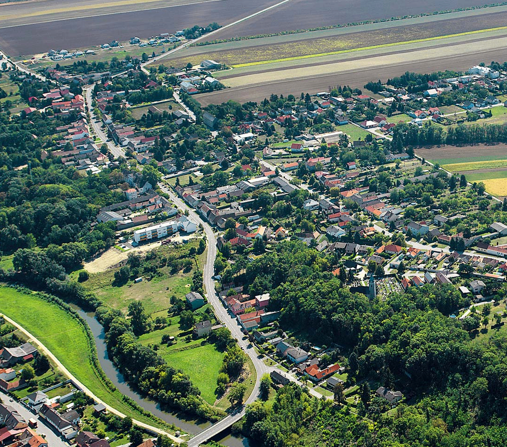  Abschnitt Bild: Eine Luftaufnahme einer Stadt mit vielen Häusern, Bäumen und einem Fluss. Die Stadt ist von Ackerland umgeben, und eine Straße verläuft durch die Mitte.